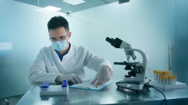 Laboratuvar tüpleriyle koruyucu tıbbi maske takan bir bilim adamı. İlerici bilimsel biyoteknoloji tıp laboratuvarı. Modern tıbbi araştırma laboratuvarında mikroskop altında araştıran erkek bilim adamı