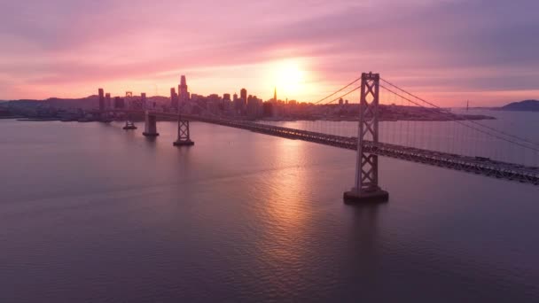 沿着海湾大桥的建筑在水面上飞行的无人机 场景景观4K 旧金山市中心有摩天大楼的海湾大桥的美丽的空中景观 在电影般的玫瑰金色落日的光芒下 — 图库视频影像