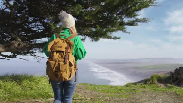 バックパックと幸せな観光の女性の風景と緑の丘を歩いてのバックビューポイントレイビーチ北カリフォルニア 米国での自然休暇中に赤カメラで撮影4Kスローモーション — ストック動画