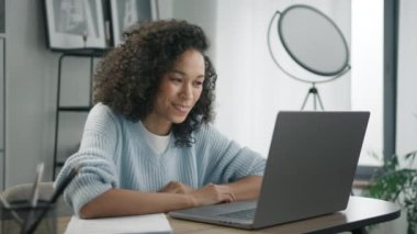 Gülümseyen Afrikalı kız öğrenci online öğretmenle e-öğrenim yapıyor, öğretmenle konferans görüşmesi yapıyor. Videolu sohbet öğrenen kız notlar alıyor, ofis masasına oturuyor, dizüstü bilgisayarına bakıyor.