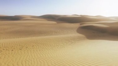 İnsansız hava aracı, üzerinde mavi gökyüzü olan altın kum tepelerinin üzerinde ufka doğru uçuyor. Derin gölgeli kum tepesi dokusu. Gün batımında hava manzaralı çöl doğası 4K. Güzel Sahra Çölü manzarası