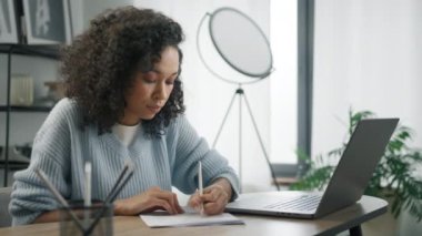 Odaklanmış ve konsantre olmuş Afrikalı kız öğrenci internet sitesinden öğreniyor. Ofis masasında uzaktan çalışan siyahi genç bir kadın, iş belgelerine not alıyor, dizüstü bilgisayarına bakıyor.