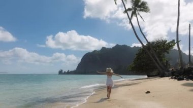 Cennet adası Oahu 'da yaz tatilindeki yavaş çekim kadın turist. Beyaz mini boho elbiseli kadın yeşil Hawaii adalarında, USA 4K. Tropikal kumsalda yürüyen bir kadın ayak izleri bırakıyor.