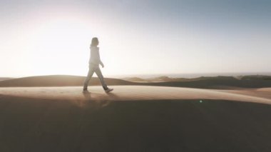 Çölde rüzgarlı bir günde beyaz kadını takip eden drone. Kum tepesinin tepesinde mutlu yürüyüşçü bir kadın mercekli altın gün batımında. Özgür kadın gezgin sinematik doğal manzara 4K 'nın yanından geçiyor.