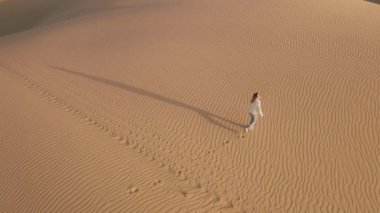 Beyaz kadın üzerinde altın ışıkta uçan drone. Sinematik kum tepeleri çölünde gezen maceraperest. Gün batımında sinemanın önünden geçen kadın gezginin yukarıdan görünüşü 4K