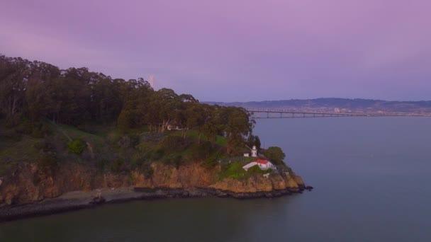 在Yerba Buena岛上的一座小型历史灯塔的空中景观 在电影的粉色紫色夕阳下 景色宜人的海湾大桥背景下的绿色宝岛上有红色屋顶的灯塔建筑 — 图库视频影像