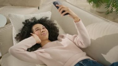 Tatillerde kapalı boş zaman. Rahat gülümseyen Afro-Amerikan kadın selfie ya da video çağrısı yapıyor, akıllı telefonu güzel yüzünün üstünde tutuyor modern bir dairede ya da otel odasında rahat bir kanepede yatıyor.