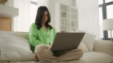 Uzaktan eğitim alan bir sınıf modern apartman dairesinde oturuyor. Genç esmer kadın üniversite öğrencisi dizüstü bilgisayarla öğreniyor, notlar yazıyor, webinar veya sanal eğitim izliyor.