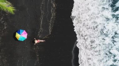 Tropikal Hawaii Adası 'nda egzotik siyah kumlu plajda oturan seksi kadın kırmızı mayokinili ve büyük hasır şapkalı günbatımı kumsalına bakıyor. Hindistan cevizi palmiyelerindeki renkli güneş şemsiyesi üzerinde tepeden aşağı hava manzarası
