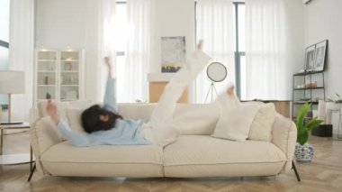 Mutluluk eğlenceli hafta sonu konsepti. Neşeli latin pozitif kadın evdeki modern oturma odasında yalın ayak kanepede zıplıyor. Mutlu kadın taşınma gününü kutluyor. Kiralık dairenin tadını çıkarın.