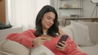 Anında kolay ödemeler yapan mutlu bayan alışverişçi, online alışveriş yapan müşteri, ve bankacılık uygulaması. Elinde akıllı telefon ve kredi kartı olan gülümseyen genç bir kadın modern evdeki koltukta oturuyor.