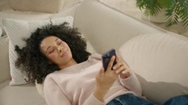 Afro saçlı mutlu genç kadın akıllı telefon kullanırken bej koltukta dinleniyor. İnternette gezinen ve arkadaşlarıyla yavaş çekimde sohbet eden çok ırklı güzel bir kadın.