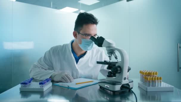 現代医学研究室で顕微鏡を見ている男性科学者 研究室の試験管で保護医療マスクをしてる科学者だ 進歩的な科学バイオテクノロジー医学研究室 — ストック動画