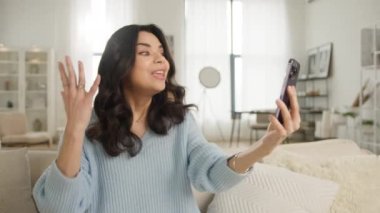 Rahat gülümseyen Latin Amerikalı kadın akıllı telefondan selfie ya da video görüşmesi yapıyor. Dairede ya da otel odasında rahat bir koltukta oturan güzel yüzlü kız. İlham verici yüz ifadeleri ve el hareketleri