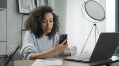 Ev ofisinde uzaktan çalışma. Üretken bir iş kadını masada oturmuş bilgisayarla çalışıyor ve akıllı telefon kamerasına bakarak görüntülü konuşuyor. Karışık ırk pozitif mızraklı kadın girişimci iş başında