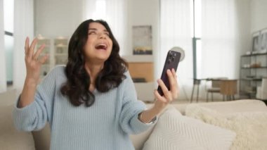 Akıllı telefondan selfie ya da video görüşmesi yapan neşeli İspanyol kadının duygusal yüz ve el hareketleri. Modern bir dairede ya da otel odası 4K 'de rahat bir koltukta oturan güzel bir kızın portresi.