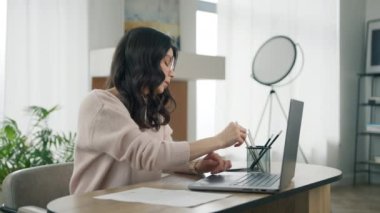 Genç İspanyol kadın üniversite öğrencisi internetten bilgisayar kullanarak öğreniyor, not alıyor, uzaktan kumandalı Webinar ya da sanal eğitim dersleri izliyor, modern apartmandaki masaüstünde oturuyor.