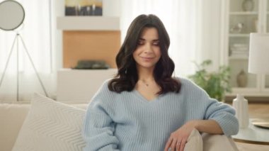 Modern oturma odasındaki dekoratif şöminenin önünde poz veren pozitif, gevşemiş İspanyol kadın. Ağır çekim millet 4K. Gülümseyen güzel Latin kadın evde tek başına oturmuş kameraya bakıyor.
