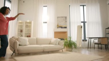 Çoklu etnik kız komik dans hareketleriyle kutlama yapıyor, hafta sonu sabah özgürlüğün tadını çıkarıyor, 4K çekim tarzı oturma odası. Yavaş hareket eden mutlu genç kadın saçlarıyla evde dans ediyor.