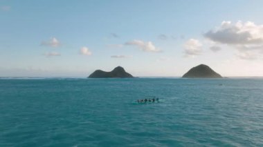 Hawaii kanoları Oahu 4K 'daki Na Mokulua adalarında denize açılıyor. Hava aracı sporcuları spor kanolarında kürek çekiyorlar. Tropikal Hawaii egzotik varış noktasında güneş doğarken turkuaz sakin bir deniz ile.