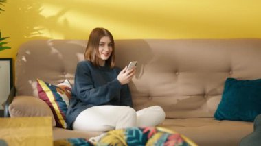 Koyu renk saçlı, kısa saçlı, akıllı telefondan yazı yazan, sohbet eden, modern parlak iç görünüşlü, kameraya gülümseyen genç bir kadın. Yüksek kalite 4k görüntü