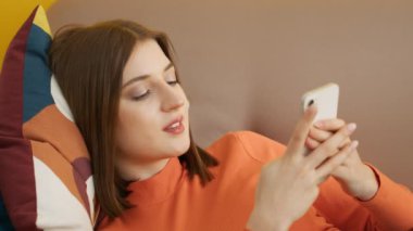 Orta boylu, neşeli bir esmer kız yan tarafta uzanıyor, akıllı telefonlara bakıyor. Kadın, başı renkli yastıkta kanepede dinleniyor. Eviniz güzelmiş. Yüksek kalite 4k görüntü