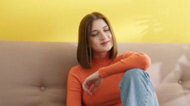 Kare saç kesimi olan çekici genç bir kadın evdeki kanepede rahatça oturuyor. Burun halkalı, hafif makyajlı, turuncu balıkçı yaka kazak ve kot pantolon giyen bir kadın. Yüksek kalite 4k görüntü