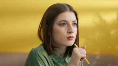 Orta boy, genç, düşünceli esmer, elinde kalem tutarken. Derin düşünceler. Mesafeye bakarak güzel bir kız öğrenci çözme problemi. Sarı arka plan. Yüksek kalite 4k görüntü
