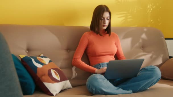 漂亮而严肃的年轻女子 深色短发 双腿交叉坐在米色沙发上 在装有黄色墙壁的房间里的银色笔记本电脑上快速打字 高质量的4K镜头 — 图库视频影像