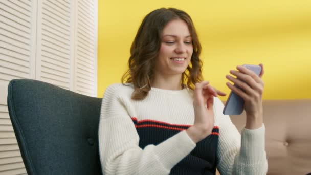 白い黒のセーターをチャットで短い巻き毛を持つ陽気な若い女性は 自宅でアームチェアに座っている 女性は携帯電話でソーシャルメディアをサーフィン リラックス 高品質4K映像 — ストック動画