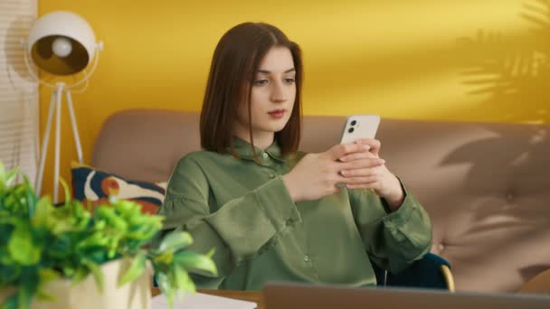 甘い顔をしたスマートなブルネットは 快適なアームチェアに座って 自宅でスマートフォンを参照してください 部屋は暖かい色で行われ 黄色の壁 ベージュのソファで 高品質4K映像 — ストック動画