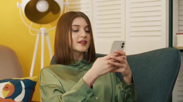 中等镜头的微笑 漂亮的黑发 浏览电话 放松在扶手椅与现代设计的房间 快乐的年轻女人聊天 在电话里打字 高质量的4K镜头 — 图库视频影像