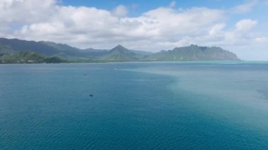 Dalış aktivitesi için mercan kayalıkları olan sığ okyanus üzerinde insansız hava aracı görüntüsü. Arka planda açık mavi suyu ve yeşil dağları olan sinematik deniz manzarası. Hawaii adasında yaz tatili