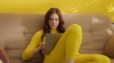 Sarıya boyanmış uzun saçlı güzel esmer bej koltukta, bacakları bükülmüş, arkasında sarı duvarlı gümüş dizüstü bilgisayarı olan bir odada oturuyor. Yüksek kalite 4k görüntü