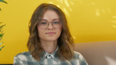 Gözlüklü, kameraya bakan orta boylu, sarışın genç bir kadın. Kısa kıvırcık saçlı, çenesi açık, koltukta oturan, sarı arka planlı bir kadın. Yüksek kalite 4k görüntü