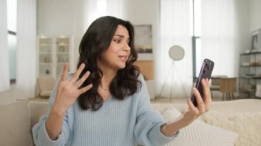 İlham verici yüz ifadeleri ve el hareketleri. Duygusal gülümseyen Latin Amerikalı kadın akıllı telefondan selfie ya da video görüşmesi yapıyor. Modern dairede rahat bir kanepede oturan güzel kız, otel odası 4K.