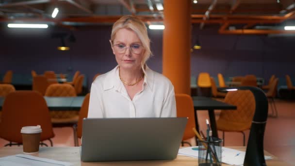 眼鏡で思慮深い焦点を当てた成熟した女性の肖像画ビュー ビジネスリーダーは 空のオフィスに座って銀製のラップトップコンピュータを見て 夜仕上げ作業に従事するご婦人上司の件 — ストック動画