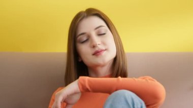Turuncu balıkçı yaka kazaklı genç bir kadın, oturuyor, gülümsüyor, el ele tutuşuyor, kısa siyah saçlarını düzeltmeye çalışıyor. Sarı arka plan. Yüksek kalite 4k görüntü