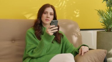 Koyu renk saçlı güzel bir kadın akıllı telefonuna bakıyor, koltukta oturuyor, kameraya baştan çıkarıcı bakışlar atıyor, gülümsüyor. Yüksek kalite 4k görüntü