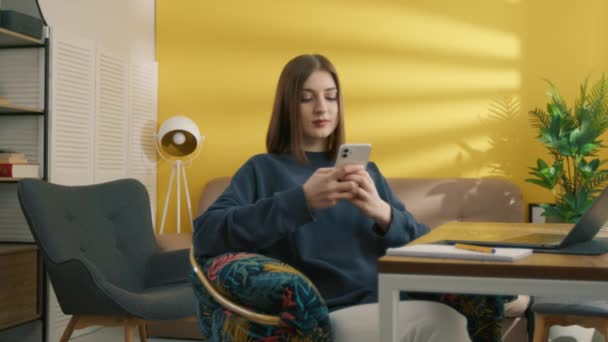 真剣に若い女性の学生は 携帯電話で入力し 彼女の明るい部屋のテーブルに座っている かなりの女性は ラップトップで勉強し 作業した後に休憩を取ります 高品質4K映像 — ストック動画