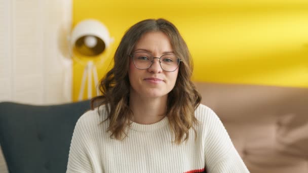 照片中带着微笑的年轻女生 戴着眼镜坐在椅子上 看着相机 房间的背景模糊不清 墙壁是黄色的 高质量的4K镜头 — 图库视频影像