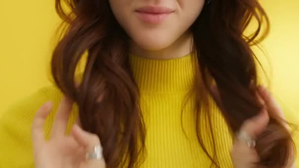 黄色の背景に黄色のセーターでわずかな笑顔で魅力的な若いブルネットの肖像画 髪に長いスタイルのカールを持つ素晴らしい女性 高品質4K映像 — ストック動画