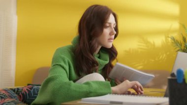 Evde dizüstü bilgisayardan bilgi alarak notlar alan sevimli bir kız öğrenci. Kadın masada oturuyor, bacağı sandalyeye sıkışmış, dizinde defteri tutuyor. Yüksek kalite 4k görüntü