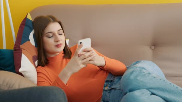 オレンジ色のタートルネックセーターとジーンズの愛の美しい若い笑顔の女性は 光の部屋でベージュのソファの上に横になり 携帯電話で高速入力します 高品質4K映像 — ストック動画