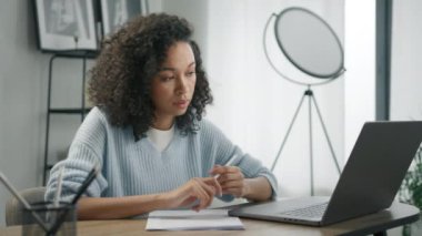 Konsantre olmuş renkli bir kadın ofis masasında uzaktan çalışıyor, iş belgelerine not alıyor, dizüstü bilgisayarına bakıyor. Odaklanmış Afrikalı kız öğrenci İnternet sitesi 4K 'de öğreniyor