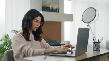 Modern ev ofisinde kahve içen odaklanmış akıllı bir kadının portresi. Çekici İspanyol bayan evde ağır çekimde kahve içiyor. Yoğun girişimci kadın gündüz dizüstü bilgisayarda çalışıyor.
