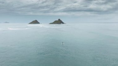 Na Mokulua adaları boyunca sörf tahtasında kürek çeken atletik erkekleri takip eden drone. Hawaii 'deki Oahu Adası' nda bulunan Kailua Körfezi 'nde. Su sporları ve bulutlu tatil gününde etkinlik, USA 4K