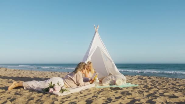 电影拍摄的母亲躺在海滩上的白色帐篷里 给小女孩读书 漂亮可爱的学龄前儿童与父母在一起很开心 年轻的金发女子为女儿创造回忆 — 图库视频影像