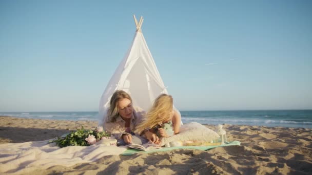 白い居心地の良い子供たちのテントで本を読んでいる幸せな家族は 日没時に海のビーチに設置され シャボン玉が飛び回っています 遊びの概念の教育 幸せなCaucasianお母さんと可愛いです赤ちゃん女の子一緒に時間を過ごす — ストック動画