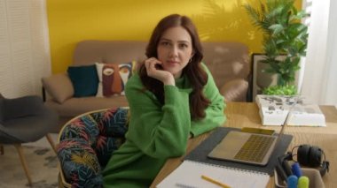 Sevimli bir kadın kameraya baştan çıkarıcı bir şekilde bakıyor, iş yerinde modern dekorla rahat bir odada oturuyor. Dizüstü bilgisayar, defter, kalem, kitap, kulaklıklar masaya. Yüksek kalite 4k görüntü